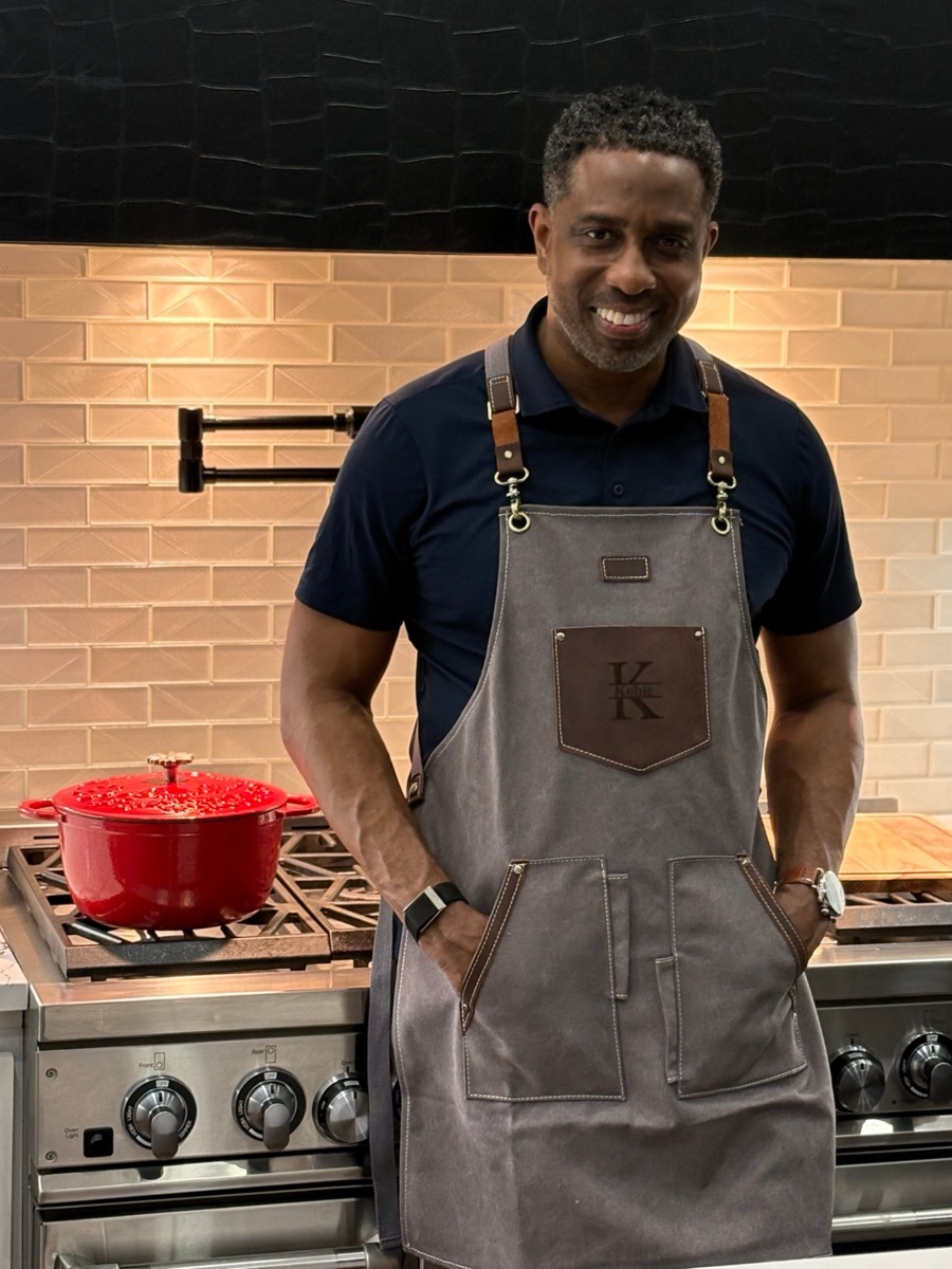 Kobie Baker in an apron at the stove, smiling, with a red Dutch oven on the burner